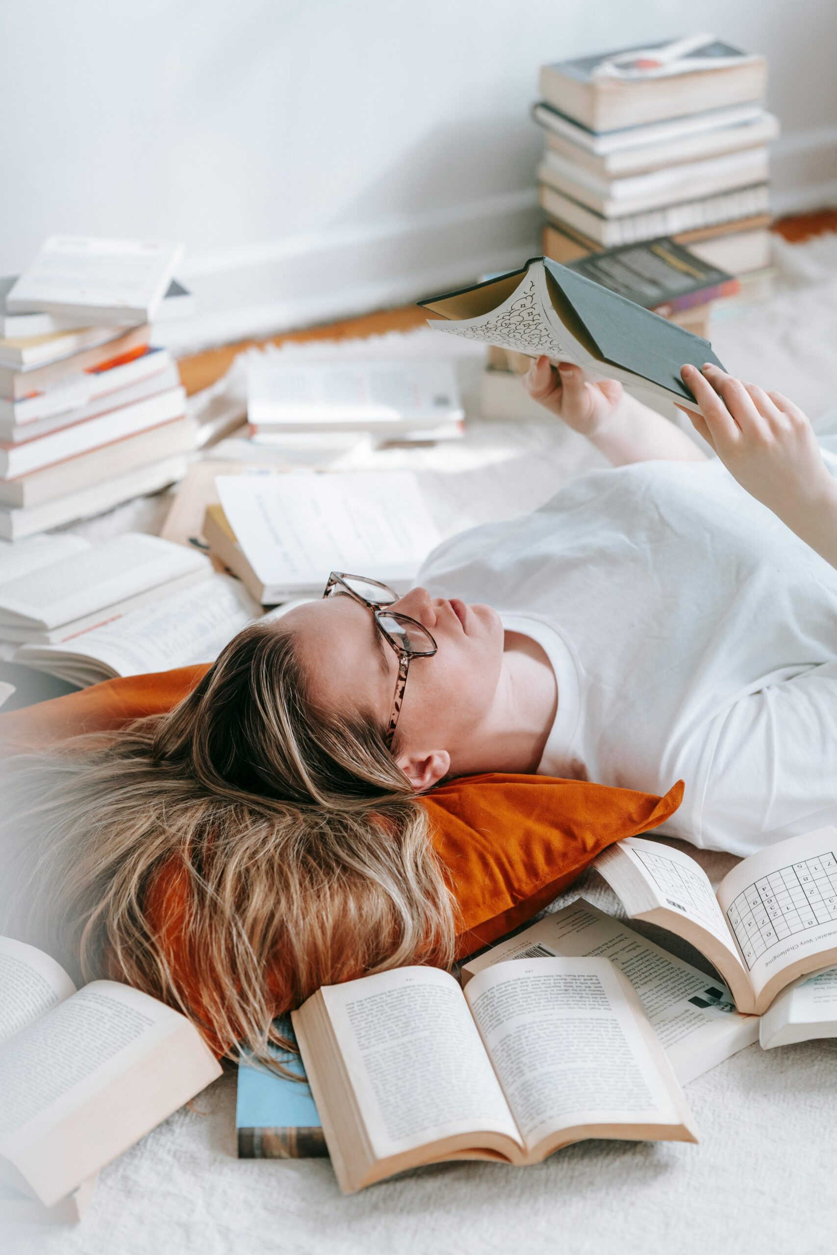 From above of calm female in eyeglasses lying on pillow and reading novel while spending time in light room with abundance of literature