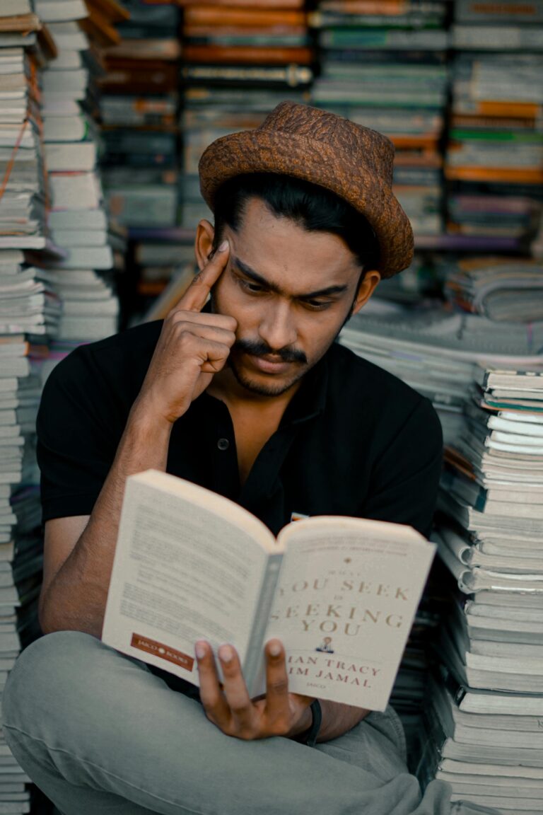 A thoughtful man in a hat reads a book in a bookstore surrounded by stacks of books.